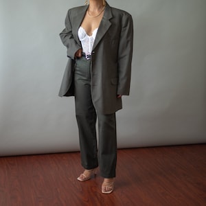 Könnte beinhalten: Ein dunkelgrüner Blazer mit langer, lockerer Passform. Der Blazer hat einen einreihigen Verschluss und einen Schalkragen. Das Model trägt den Blazer über einem weißen Spitzenoberteil und einer dunkelgrünen Hose.