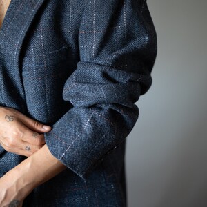 Op de afbeelding: Een close-up van een persoon die een donkerblauwe blazer draagt met een subtiel herringbone-patroon. De blazer heeft een dubbele rij knopen en een enkele knoop op elke manchet.