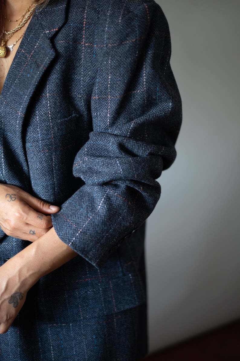 Op de afbeelding: Een close-up van een persoon die een donkerblauwe blazer draagt met een subtiel herringbone-patroon. De blazer heeft een dubbele rij knopen en een enkele knoop op elke manchet.