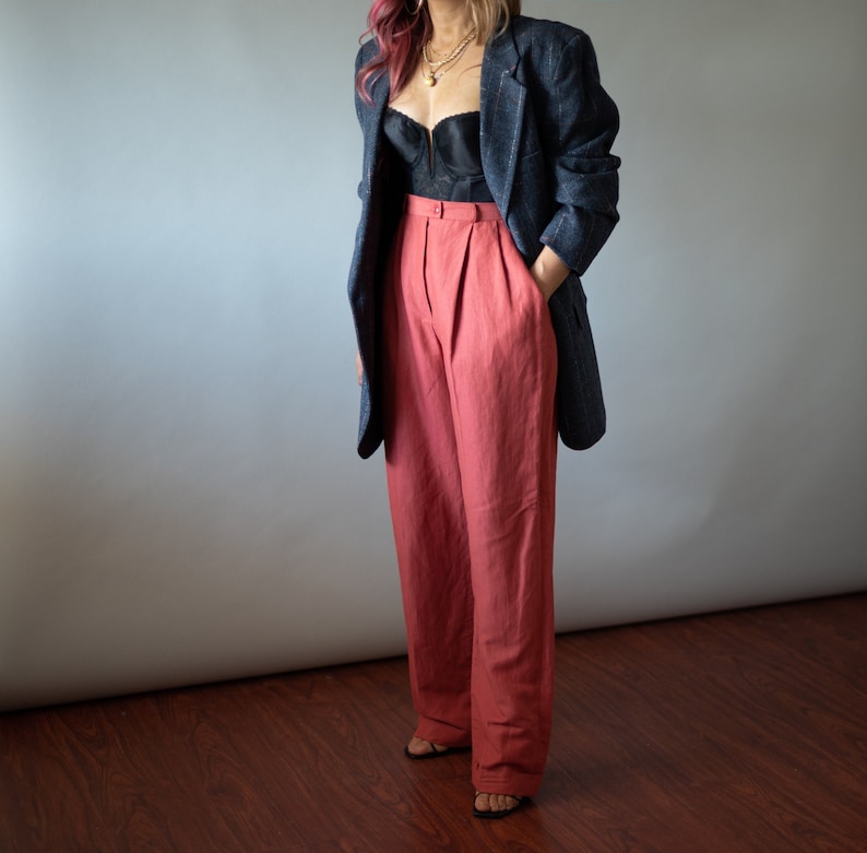 Op de afbeelding: Een vrouw draagt een zwart blazer met een ruitpatroon over een zwart kanten top en een wijde, koraal gekleurde broek.