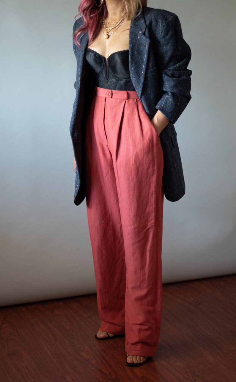 Op de afbeelding: Een vrouw draagt een zwarte kanten bustier, een donkerblauw blazer en een wijde, roestkleurige broek. Ze draagt gouden kettingen en heeft roze haar.