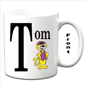 May include: White ceramic mug with a black letter "T" and the word "Tom" printed on the front. A cartoon yellow cat wearing a purple waistcoat and a hat is next to the text. The word "Front" is printed on the handle.