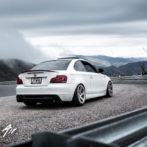 K&ouml;nnte beinhalten: Ein wei&szlig;er BMW 1er Coup&eacute; f&auml;hrt auf einer kurvigen Stra&szlig;e mit Blick auf die Berge im Hintergrund. Das Auto hat einen schwarzen Heckspoiler und ein Nummernschild mit der Aufschrift "JTW56".