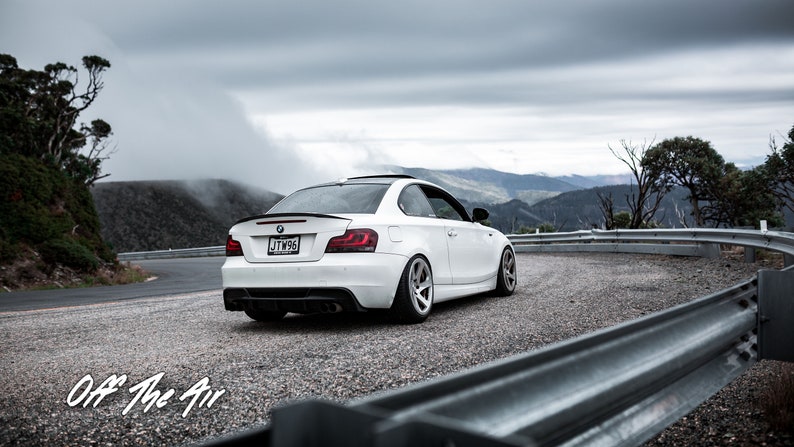K&ouml;nnte beinhalten: Ein wei&szlig;er BMW 1er Coup&eacute; f&auml;hrt auf einer kurvigen Stra&szlig;e mit Blick auf die Berge im Hintergrund. Das Auto hat einen schwarzen Heckspoiler und ein Nummernschild mit der Aufschrift "JTW56".