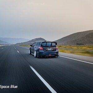 May include: A dark blue Subaru WRX STI driving on a highway with mountains in the background. The car has a rear spoiler and a license plate that reads "WRX STI".