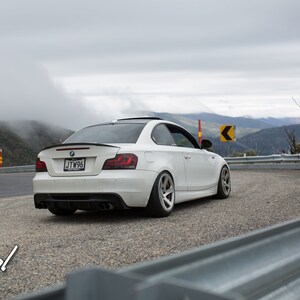 K&ouml;nnte beinhalten: Ein wei&szlig;er BMW 1er Coup&eacute;, das auf einer kurvigen Stra&szlig;e mit Blick auf die Berge im Hintergrund geparkt ist. Das Auto hat einen schwarzen Spoiler und schwarze R&auml;der. Das Kennzeichen lautet "JTW96".