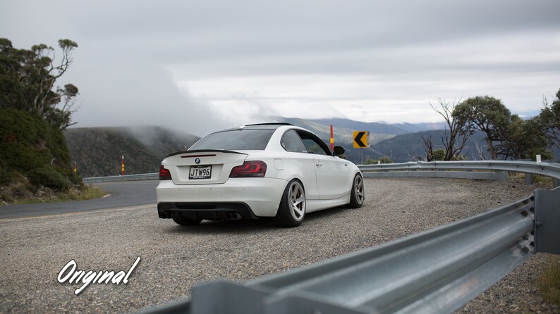 K&ouml;nnte beinhalten: Ein wei&szlig;er BMW 1er Coup&eacute;, das auf einer kurvigen Stra&szlig;e mit Blick auf die Berge im Hintergrund geparkt ist. Das Auto hat einen schwarzen Spoiler und schwarze R&auml;der. Das Kennzeichen lautet "JTW96".