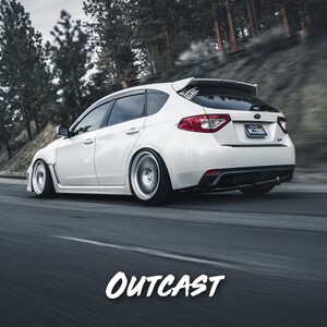 K&ouml;nnte beinhalten: Ein wei&szlig;er Subaru Impreza WRX STI Kombi f&auml;hrt auf einer kurvigen Stra&szlig;e durch einen Wald. Das Auto hat einen schwarzen Heckspoiler und schwarze Felgen. Das Auto ist in Bewegung und der Hintergrund ist verschwommen.