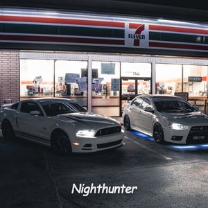 May include: Two white sports cars parked in front of a convenience store at night. The car on the left is a Ford Mustang and the car on the right is a Mitsubishi Lancer Evolution. Both cars have aftermarket LED lights under the body.