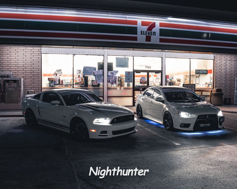 May include: Two white sports cars parked in front of a convenience store at night. The car on the left is a Ford Mustang and the car on the right is a Mitsubishi Lancer Evolution. Both cars have aftermarket LED lights under the body.