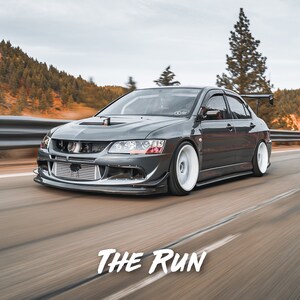 K&ouml;nnte beinhalten: Ein grauer Mitsubishi Lancer Evolution rast auf einer Autobahn mit einer Bergkette im Hintergrund. Das Auto hat einen gro&szlig;en Heckfl&uuml;gel und wei&szlig;e R&auml;der. Der Text "THE RUN" ist im unteren mittleren Bereich des Bildes sichtbar.