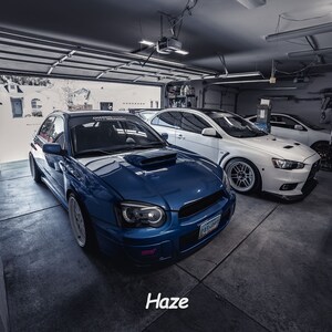 May include: A blue Subaru Impreza WRX STI with a white racing stripe and black aftermarket headlights parked in a garage with a white Mitsubishi Lancer Evolution parked behind it.
