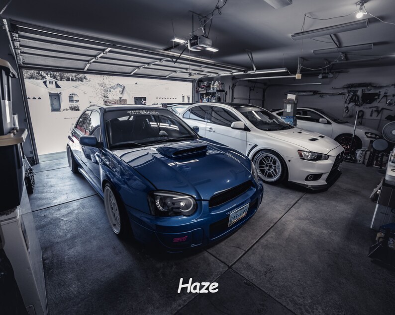 May include: A blue Subaru Impreza WRX STI with a white racing stripe and black aftermarket headlights parked in a garage with a white Mitsubishi Lancer Evolution parked behind it.