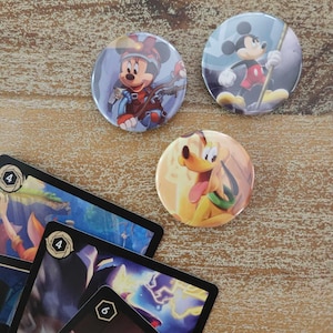 May include: Three round pinback buttons featuring Disney characters. The buttons are on a wooden surface. The first button features Mickey Mouse in a red and yellow outfit climbing a rock face. The second button features Mickey Mouse in a red and yellow outfit holding a staff. The third button features Pluto in a yellow and green outfit.