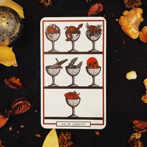 May include: A tarot card with a white background and black lines. The card depicts seven goblets with various objects inside, including a skull, a heart, a snake, and a sword. The card is titled "VII of Goblets".