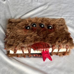 May include: A rectangular book covered in brown faux fur featuring four black spherical eyes on the front cover. The edges of the book are adorned with brown sculpted claws and a central red mouth with sharp teeth and a protruding red forked tongue.