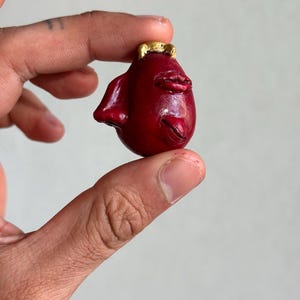 May include: A small, dark red, rounded sculpture with a smooth, glossy finish. It features a prominent, textured pair of lips, a small pointed ear on the side, and a metallic gold-colored accent at the top, resembling a small crown or horn.