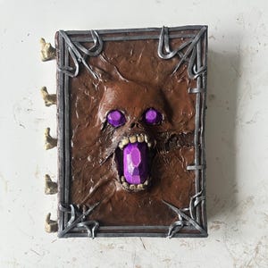 May include: A brown leather-bound book with a skeletal face and purple gem on the cover. The book has metal clasps and bone-like page holders.
