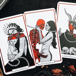 May include: Three tarot cards with black and white illustrations and red accents. The Empress card depicts a woman holding a baby, The Lovers card shows a skeleton embracing a woman, and The Devil card features a horned figure with a skull face.