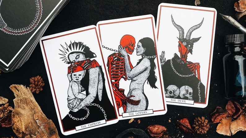 May include: Three tarot cards with black and white illustrations and red accents. The Empress card depicts a woman holding a baby, The Lovers card shows a skeleton embracing a woman, and The Devil card features a horned figure with a skull face.
