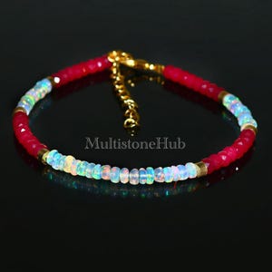 May include: A delicate bracelet featuring alternating red and white beads. The white beads are iridescent and have a rainbow effect. The bracelet has a gold clasp.