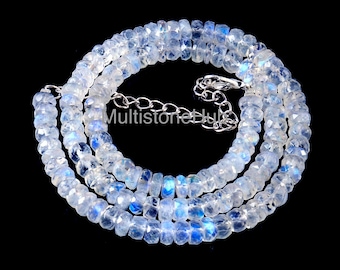 Op de afbeelding: Een ketting van gefacetteerde, doorschijnende maansteen kralen met blauwe en witte flitsen. De kralen zijn geregen met een zilverkleurige ketting en sluiting. De ketting wordt gepresenteerd tegen een zwarte achtergrond.