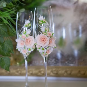 Op de afbeelding: Twee champagnefluiten versierd met delicate roze en witte bloemen. De bloemen zijn gemaakt van stof en zijn gerangschikt in een tros rond de bovenkant van elke fluit.
