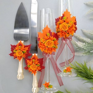 May include: A wedding cake cutting set with two champagne flutes decorated with orange and red fall leaves and flowers. The set includes a knife and a server with white handles.