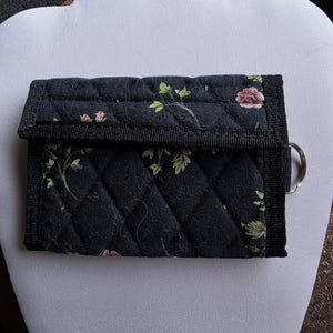 May include: A black quilted tri-fold wallet featuring a floral pattern with small pink flowers and green leaves. The wallet has a black fabric trim, a fold-over flap closure, and a silver-toned metal ring attached to the side for key attachment.
