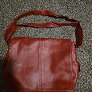 Könnte beinhalten: Eine rote Messenger-Tasche aus Leder mit einem langen Schulterriemen. Die Tasche hat einen Klappverschluss und zwei Seitentaschen. Die Tasche besteht aus einem glatten, glänzenden Material und hat ein genähtes Design in der Mitte.