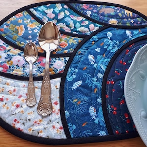 May include: A quilted placemat with a black border, featuring sections of floral and marine-themed fabrics in blue, teal, and white. Two silver spoons and two decorative plates are on the placemat. The placemat is on a wooden surface.