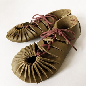 May include: A pair of olive green leather shoes featuring a gathered, pleated toe design and a lace-up front closure with thin, dark red cord laces. The shoes have a low-cut ankle profile and a smooth, matte finish on the exterior leather surface.