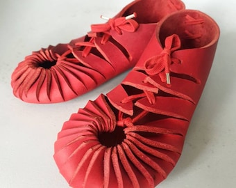 May include: A pair of red leather sandals with a unique, pleated design around the toe. The sandals have laces that tie at the ankle.
