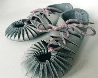 May include: A pair of gray leather shoes with a unique pleated design around the toe and a lace-up closure. The laces are a muted pink color. The shoes are displayed on a white background.