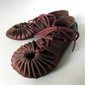 Könnte beinhalten: Ein Paar handgefertigte, dunkelburgunderfarbene Lederschuhe. Die Schuhe haben ein einzigartiges Design mit plissiertem Leder um die Zehen und burgunderfarbenen Schnürsenkeln. Die Schuhe scheinen ungetragen zu sein und betonen die Handwerkskunst. Ca. 25 cm lang.