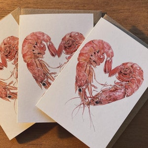 Three small cards each featuring a design of 2 shrimp curled around to form a heart shape. They are placed on a wooden background, and there are brown envelopes inside each of the cards.