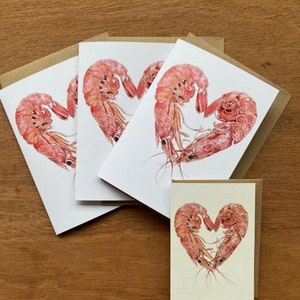 3 larger folded cards featuring heart shaped prawns lie on a plain wooden background. A smaller version of the same card is at the bottom right hand of the image.