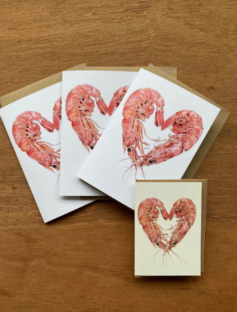 3 larger folded cards featuring heart shaped prawns lie on a plain wooden background. A smaller version of the same card is at the bottom right hand of the image.