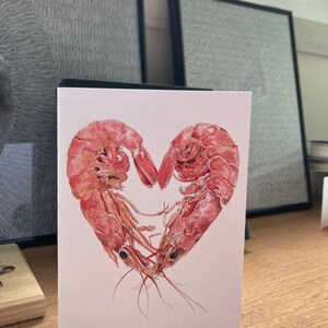 A card with 2 pink prawns curled round each other to form a heart shape on a plain white background. In the background are 2 framed drawings.