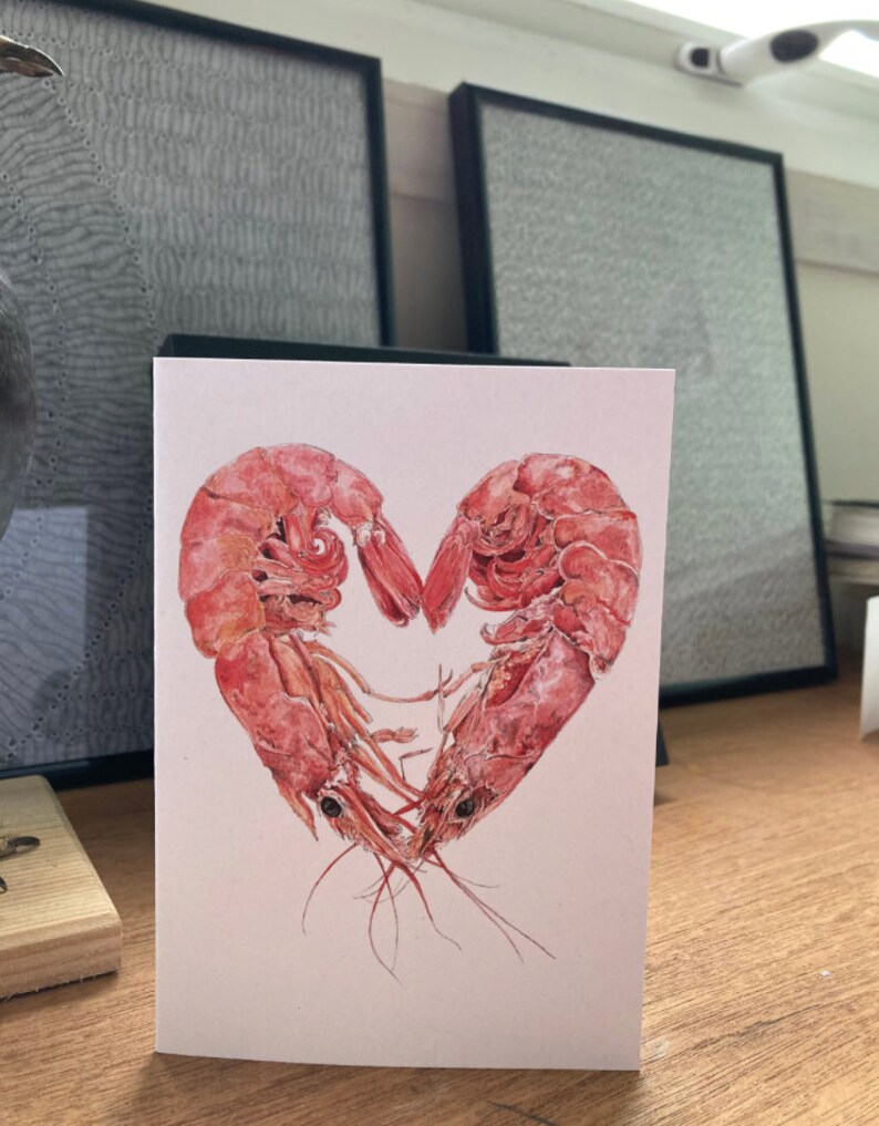 A card with 2 pink prawns curled round each other to form a heart shape on a plain white background. In the background are 2 framed drawings.