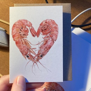 A hand with unpainted nails holds a small card with 2 prawns curled together to form a heart shape.