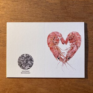An unfolded card is placed on a plain wooden background. The card shows both the front and back. The front shows 2 prawns curled round to form a heart, and the back shows a monochrome logo.