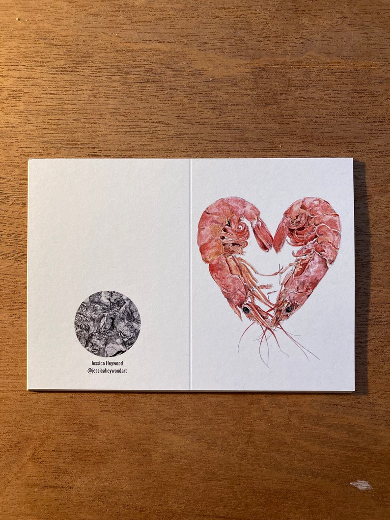 An unfolded card is placed on a plain wooden background. The card shows both the front and back. The front shows 2 prawns curled round to form a heart, and the back shows a monochrome logo.