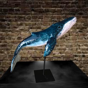 Pode incluir: Uma escultura de baleia azul e branca montada em um suporte de metal preto. A baleia é retratada em uma pose dinâmica, com o corpo arqueado. A escultura tem um acabamento brilhante, e os detalhes da forma da baleia são claramente visíveis.