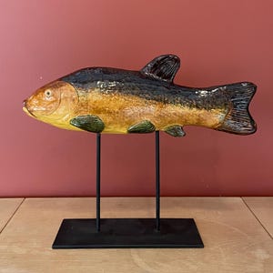 Könnte beinhalten: Eine Keramik-Fischskulptur mit schwarzer, brauner und gelber Glasur. Der Fisch ist auf einem schwarzen Metallständer mit zwei Beinen montiert.