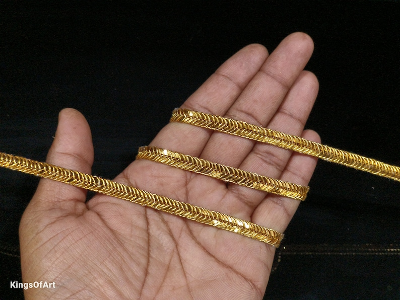 May include: Gold trim with a zig-zag pattern. The trim is shown on a hand, with three strands of the trim.