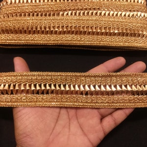 May include: Gold-colored decorative trim with a textured, woven design. The trim features a repeating pattern of triangular shapes along one edge. The trim is likely used for embellishing clothing or craft projects.