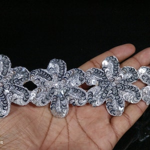 Puede incluir: Borde de lentejuelas plateadas con un diseño floral. El borde está hecho de pequeñas lentejuelas plateadas cosidas sobre una base de tela negra.