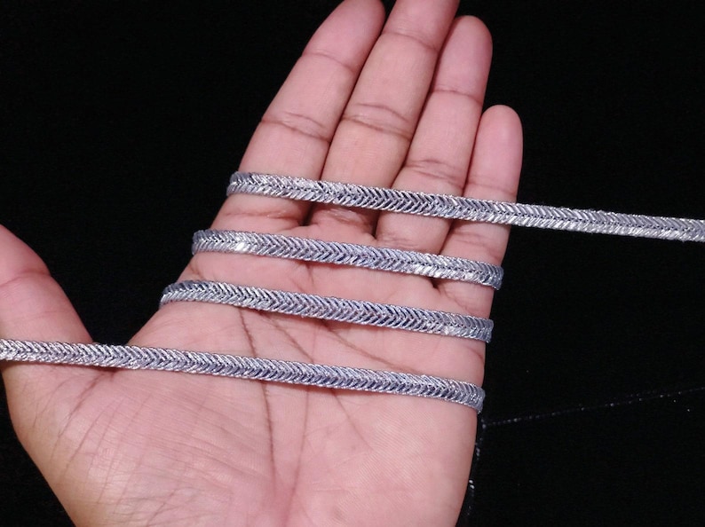 May include: Silver trim with a zig-zag pattern. The trim is shown in a hand, folded over on itself.