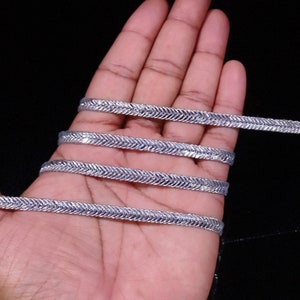 May include: Silver trim with a herringbone pattern. The trim is shown in a hand, with multiple strands of the trim.